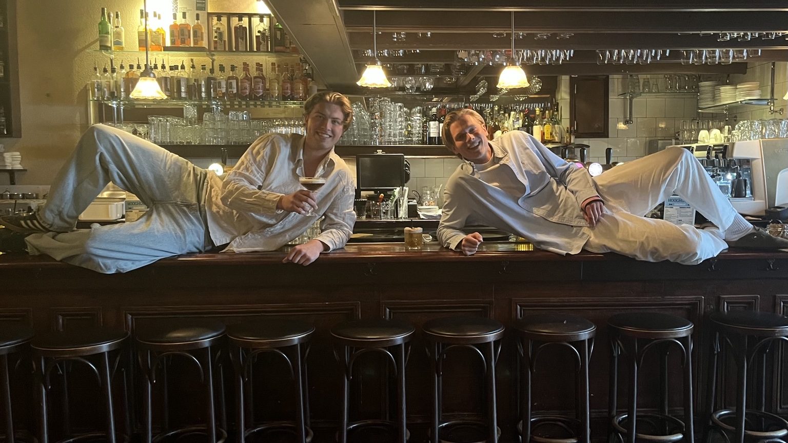 Karsten en Coen liggen lachend op de bar, met een cocktail binnen handbereik.